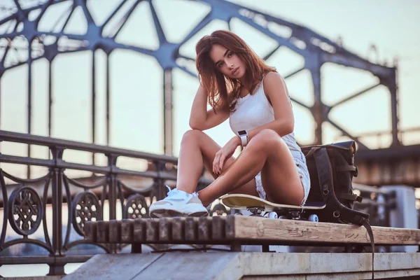 Jovem sensual skater menina vestida com shorts e t-shirt sentado no dique durante belo pôr do sol . — Fotografia de Stock