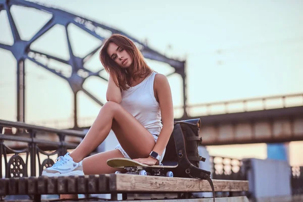 ショート パンツや美しい日没時に堤防の上に座って t シャツに身を包んだ若い官能的なスケーターの女の子. — ストック写真