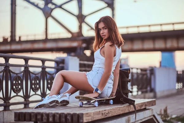 Jovem sensual skater menina vestida com shorts e t-shirt sentado no dique durante belo pôr do sol . — Fotografia de Stock