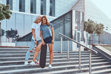 Bir iki genç hippi kız gülümseyerek portresi adımları gökdelen bir arka plan üzerinde duran bir kaykay tutar. 