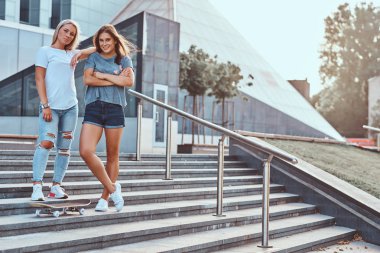 İki güzel hipster kızlar merdivenlerinde gökdelen bir arka plan üzerinde kaykay ile daimi.