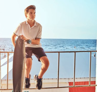 Bir genç kaykaycı çocuk portresi t-shirt ve şort Otokorkuluk üzerinde oturan giymiş ve bir kaykay sahil arka planı tutar.