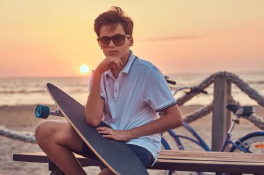 T-shirt ve şort parlak günbatımında bir sahil çerçevede bir bankta oturan genç dalgın patenci çocuk güneş gözlüğü giymiş.