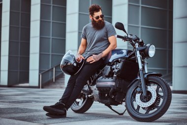 Gri tişört ve siyah pantolon acımasız bir sakallı erkek portresi bir gökdelen karşı özel yapım retro motosiklet üzerinde oturan bir kask tutar