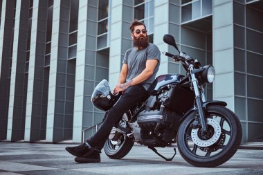 Gri tişört ve siyah pantolon acımasız bir sakallı erkek portresi bir gökdelen karşı özel yapım retro motosiklet üzerinde oturan bir kask tutar