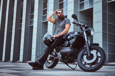 Giyinmiş bardaklarda acımasız sakallı erkek gri tişört ve siyah pantolon saç düzeltmek ve bir gökdelen karşı özel yapım retro motosiklet üzerinde oturan bir kask tutar.