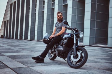 Acımasız sakallı erkek gri tişört ve siyah pantolon bir gökdelen karşı özel yapım retro motosiklet üzerinde oturan bir kask tutar.