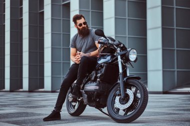 Gri tişört ve siyah pantolon gökdelen karşı özel yapım retro motosiklet üzerinde oturan acımasız sakallı erkek güneş gözlüğü giymiş.