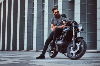 Gri tişört ve siyah pantolon gökdelen karşı özel yapım retro motosiklet üzerinde oturan acımasız sakallı erkek güneş gözlüğü giymiş.