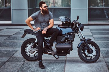 Gri tişört ve siyah pantolon gökdelen karşı özel yapım retro motosiklet üzerinde oturan acımasız sakallı erkek güneş gözlüğü giymiş.