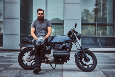 Bir kameraya bakarak bir gökdelen karşı özel yapım retro motosiklet üzerinde oturan bir kask Gri tişört ve siyah pantolon acımasız sakallı erkek tutar.