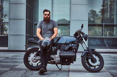 Bir kameraya bakarak bir gökdelen karşı özel yapım retro motosiklet üzerinde oturan bir kask Gri tişört ve siyah pantolon acımasız sakallı erkek tutar.