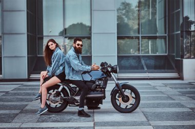 Çekici hipster çift - sakallı vahşi erkek güneş gözlüğü ve kot ceket ve arka arkaya bir gökdelen karşı retro bir motosiklet üzerinde oturan genç onun şehvetli kız.