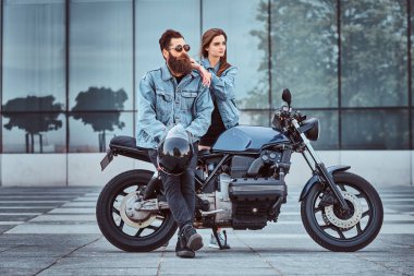 Retro bir motosiklet ve yakın duran, gökdelen karşı poz genç onun şehvetli kız oturan çekici hipster çift - sakallı vahşi erkek güneş gözlüğü ve kot pantolon ceket.