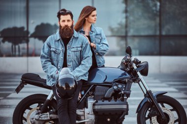 Retro bir motosiklet ve yakın duran, gökdelen karşı poz genç onun şehvetli kız oturan çekici hipster çift - sakallı vahşi erkek güneş gözlüğü ve kot pantolon ceket.