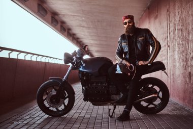 Şık bir sakallı erkek portresi bir kameraya bakarak yaptığı ısmarlama retro motosiklet üzerinde oturan gözlüklü bir siyah deri ceket ve bandana giymiş.