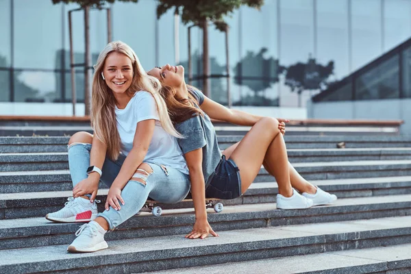 高層ビルの背景にステップでスケート ボードに一緒に座っている二人の流行に敏感な若い女の子の肖像画 — ストック写真