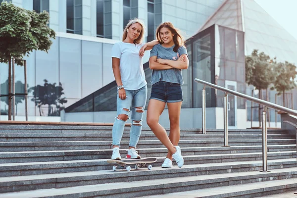 Zwei schöne Hipstermädchen, die auf Stufen mit Skateboard vor dem Hintergrund des Wolkenkratzers stehen. — Stockfoto