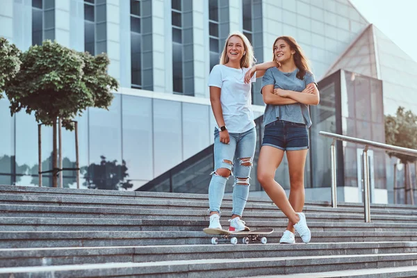 Dos hermosas chicas hipster de pie en las escaleras con monopatín sobre un fondo del rascacielos . —  Fotos de Stock