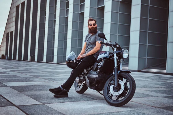 在灰色 T恤衫和黑色长裤的野蛮胡子男性持有头盔坐在他的定制复古摩托车对摩天大楼 — 图库照片