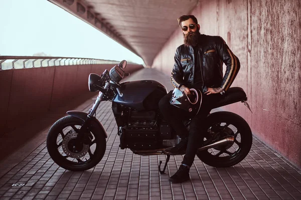 サングラスと黒い革のジャケットに身を包んだスタイリッシュなおしゃれなバイクを保持彼の特注のレトロなバイクの上に座ってヘルメット カメラを見て — ストック写真