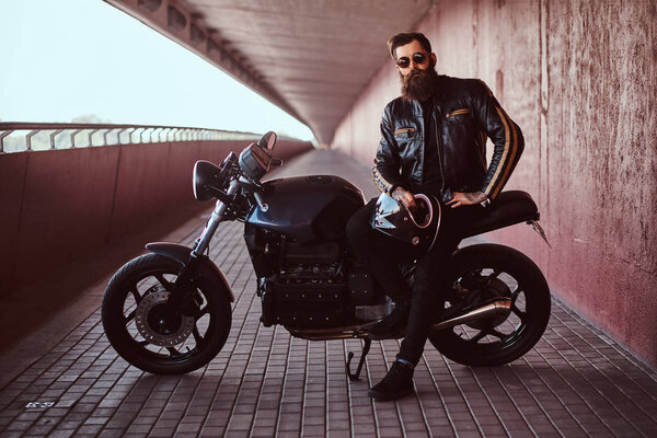 Stylish fashionable biker dressed in a black leather jacket with sunglasses holds a helmet sitting on his custom-made retro motorcycle, looking at a camera.