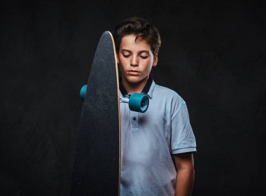 Beyaz bir t-shirt giymiş bir genç kaykaycı çocuk portresi bir longboard tutar. Karanlık bir arka plan üzerinde izole.