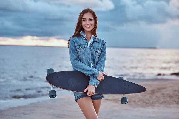 幸せの官能的なブルネットの少女は デニムのショート パンツに身を包んだし ジャケットは ビーチで立ちながらスケート ボードを保持曇りの天候で日没時に — ストック写真