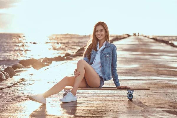 ショート パンツに身を包んだ女の子を魅力的な シャツは明るい日没時にビーチでスケート ボードに座って幸せ — ストック写真