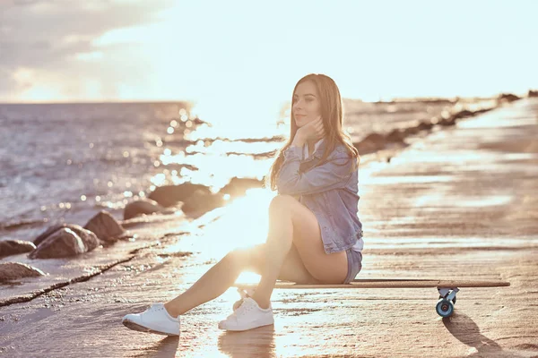 短パン姿の美しいブルネットの少女 シャツは明るい日没時にビーチでスケート ボードに座っています — ストック写真