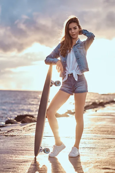 Krásná Dívka Sobě Džínové Kraťasy Sako Pózuje Skateboard Pláži Oblačné — Stock fotografie