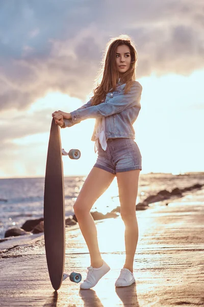 デニムのショート パンツと明るい日没時に曇りの天気でビーチでスケート ボードでポーズのジャケットに身を包んだ美しい少女 — ストック写真