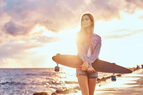 穿着牛仔短裤和夹克的性感黑发女孩在晴朗的日落时 站在海滩上 在多云的天气里举行滑板 — 图库照片