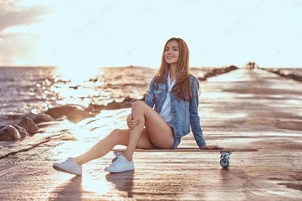 Happy charming girl dressed in shorts and a t-shirt is sitting on a skateboard on the beach during bright sunset.