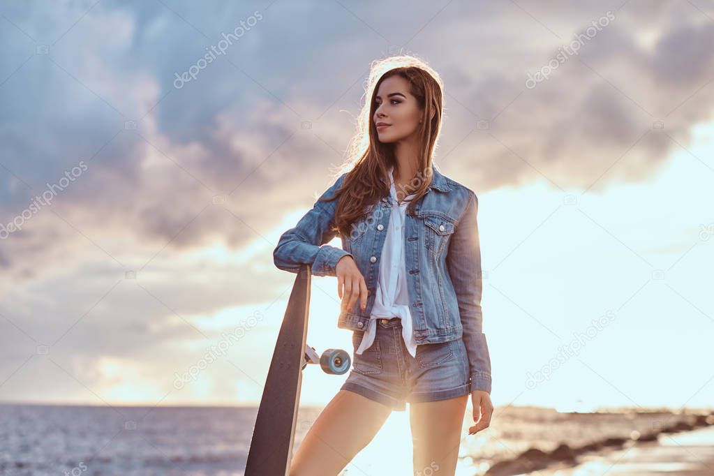 Beautiful girl dressed in denim shorts and jacket posing with a skateboard on the beach in cloudy weather during bright sunset.