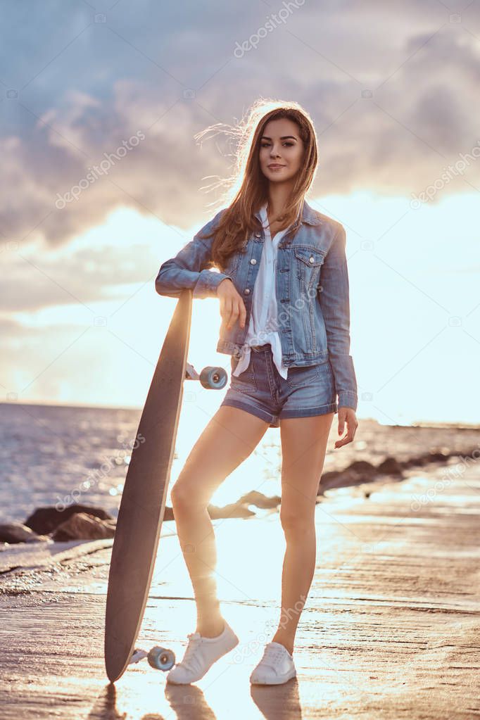 Beautiful girl dressed in denim shorts and jacket posing with a skateboard on the beach in cloudy weather during bright sunset.