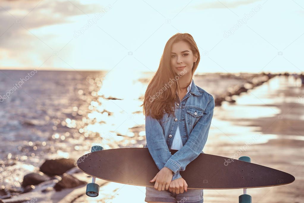 Sensual brunette girl dressed in denim shorts and jacket holds skateboard while standing on the beach in cloudy weather during bright sunset.