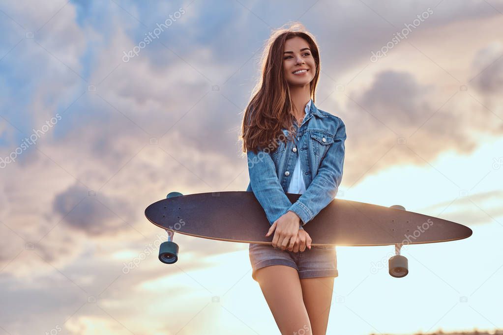 Smiling sensual brunette girl dressed in denim shorts and jacket holds skateboard while standing on the beach in cloudy weather during bright sunset.