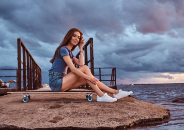 Sahilde bir kaykay üzerinde oturan gülümseyen sevimli kız inanılmaz karanlık bulutlu hava günbatımı sırasında keyif alıyor.