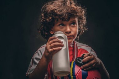 Little champion boy in sportswear holds a ball and drinking water from a bottle. Isolated on the dark textured background. clipart