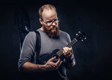 Kızıl saçlı gözlüklü bir ukulele üzerinde oynarken Gri tişört giymiş erkek müzisyen sakallı. Karanlık dokulu arka plan üzerinde izole. 
