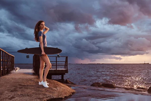 浜に立っては日没時に驚くほど暗い曇り天気を楽しんでいる間に官能的な女の子の全身肖像画がスケート ボードを保持します — ストック写真