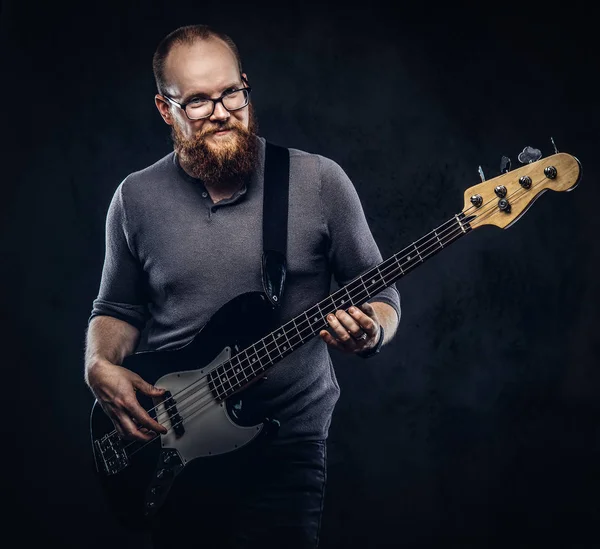 Smiling redhead bearded male musician wearing glasses dressed in a gray t-shirt playing on electric guitar. Isolated on a dark textured background.