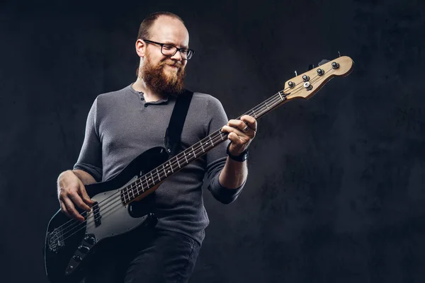 Redhead bearded male musician wearing glasses dressed in a gray t-shirt enjoying playing on electric guitar. Isolated on a dark textured background.