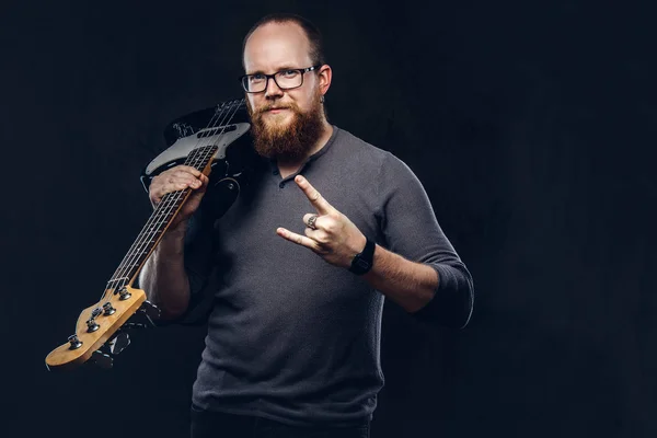 Redhead bearded male musician wearing glasses dressed in a gray t-shirt holds electric guitar and show rock and roll sign. Isolated on dark textured background.