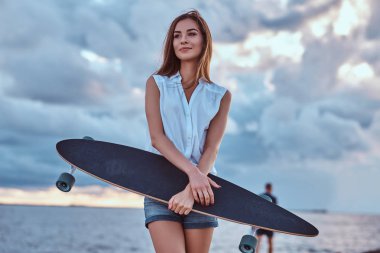 Şehvetli bir esmer kız portresi şort giymiş ve beyaz bir gömlek kaykay sahilde bulutlu havalarda günbatımı sırasında tutar.