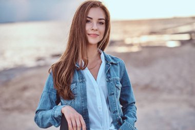 Yakın çekim portre sevimli bir kız ile bir kaykay bulutlu havalarda sahilde günbatımı sırasında poz denim ceket giymiş.