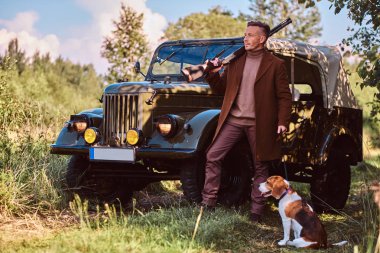Şık giysileriyle avcısı beagle köpeğini yakınındaki bir ormandaki retro bir askeri araç ile birlikte bir av tüfeği ve ayakta tutan.