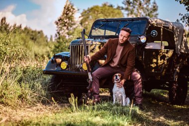 Şık giysileriyle avcı av tüfeği tutan ve retro bir askeri araç ormandaki yaslanmış iken onun beagle köpek ile birlikte oturur.