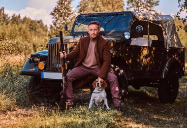 Şık giysileriyle avcı av tüfeği tutan ve retro bir askeri araç ormandaki yaslanmış iken onun beagle köpek ile birlikte oturur.